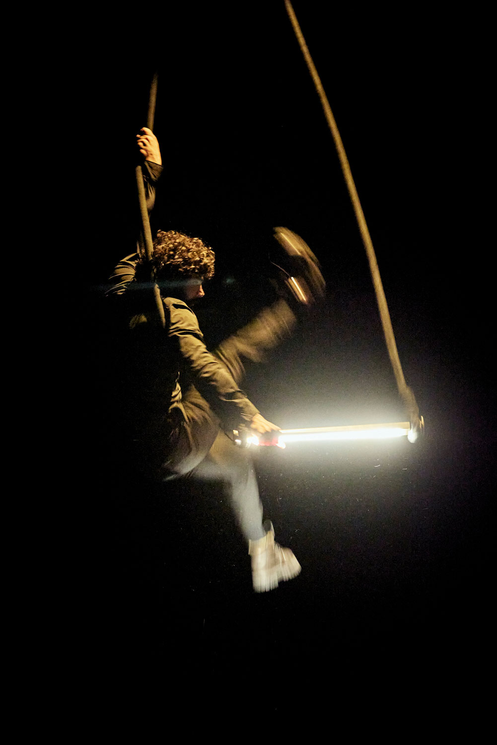 Sur scène, un circassien est assis à califourchon, sur un trapèze néon.