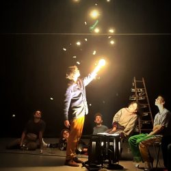 Le circassien fait voler en l'air des poussières de feuilles d'or entouré du public dans un bâtiment sombre.