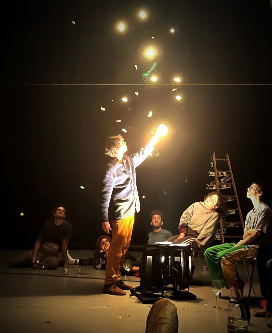 Le circassien fait voler en l'air des poussières de feuilles d'or entouré du public dans un bâtiment sombre.