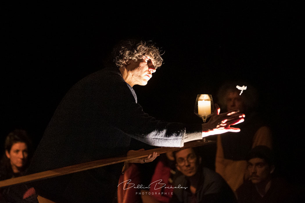 Un circassien maintient une bougie en équilibre sur une planche et est légèrement éclairé par celle-ci.