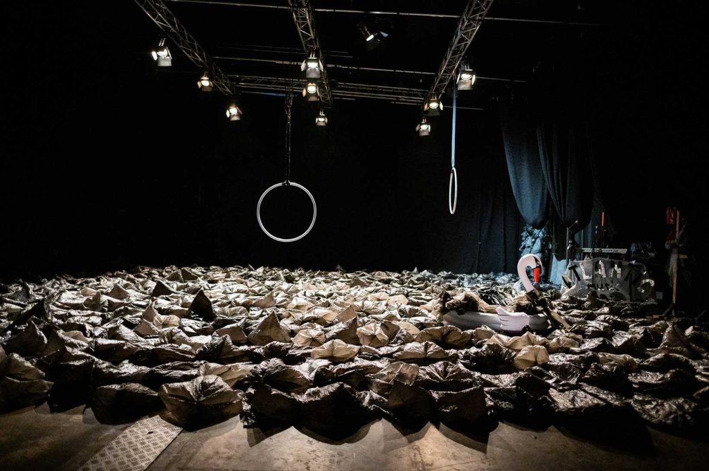 Décor de scène constitué d’un sol jonchés de sacs plastiques, 2 cerceaux suspendus en fond de scène et côté cours et un bouée cygne.