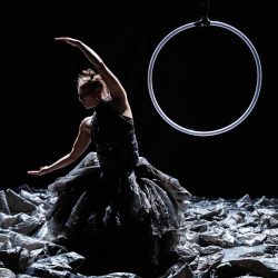 Habillée d’une de sacs plastiques une danseuse ouvre ses bras en demi-lune. Derrière elle se trouve un cerceau suspendu.