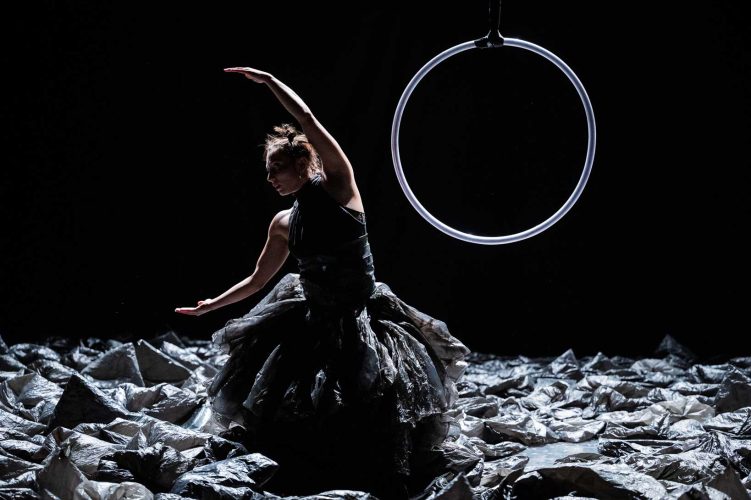 Habillée d’une de sacs plastiques une danseuse ouvre ses bras en demi-lune. Derrière elle se trouve un cerceau suspendu.