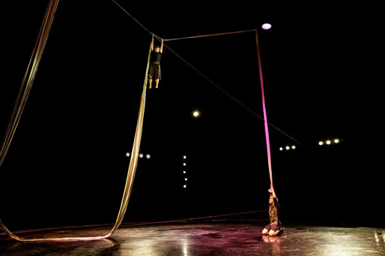 Crédit François Ollivier. En salle. Sur une scène éclairée par 2 spots, on voit un draps suspendus éclairés en jaune et rose. 2 danseuses tirent et grimpent sur ce drap en jouant avec leurs contrepoids.