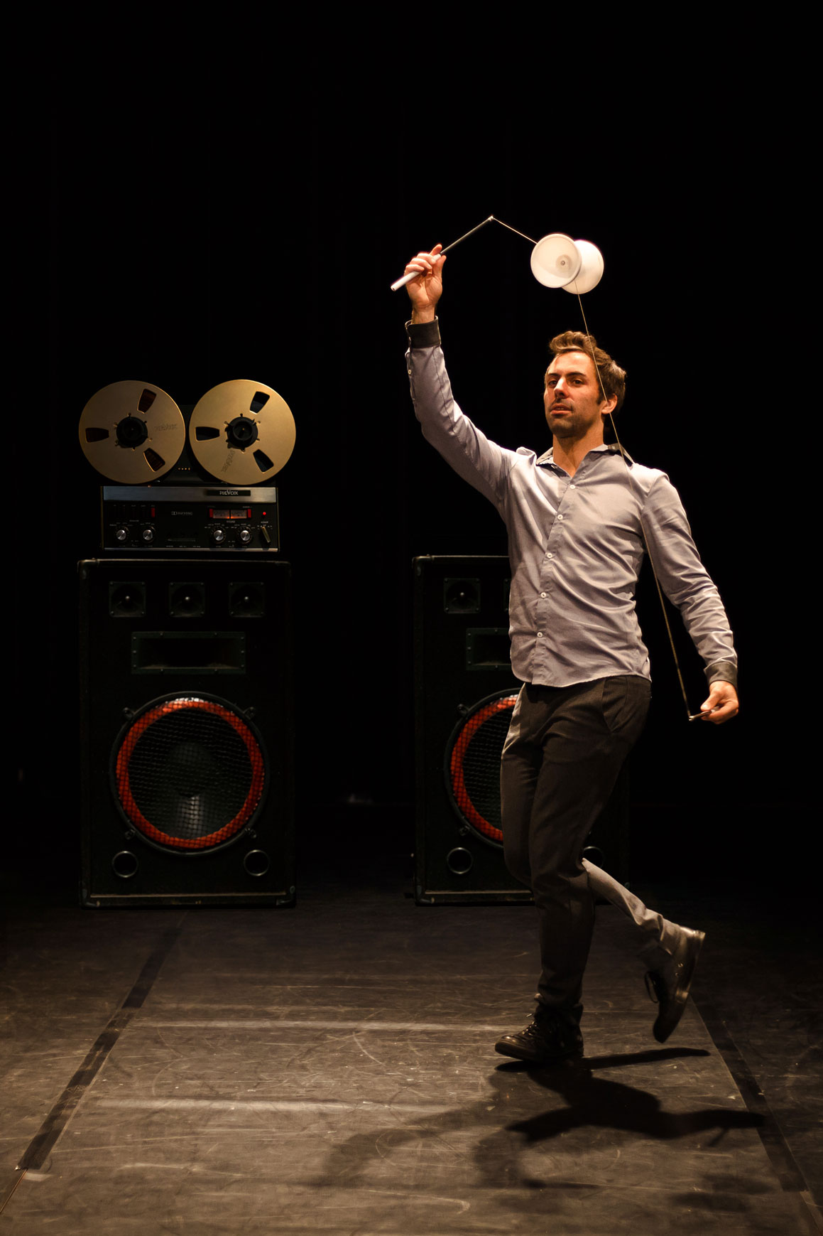 Un circassien lance un diabolo en hauteur en marchant, devant un magnétophone et deux enceintes, sous une lumière blanche.