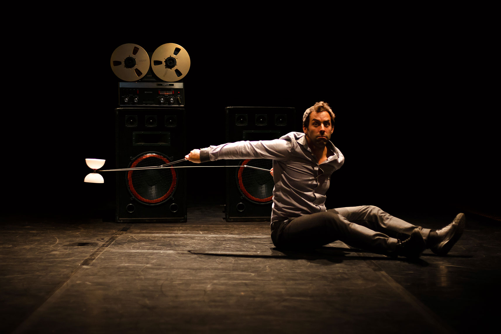 Devant un magnétophone a bande, un circassien assis au sol envoi son diabolo derrière lui.