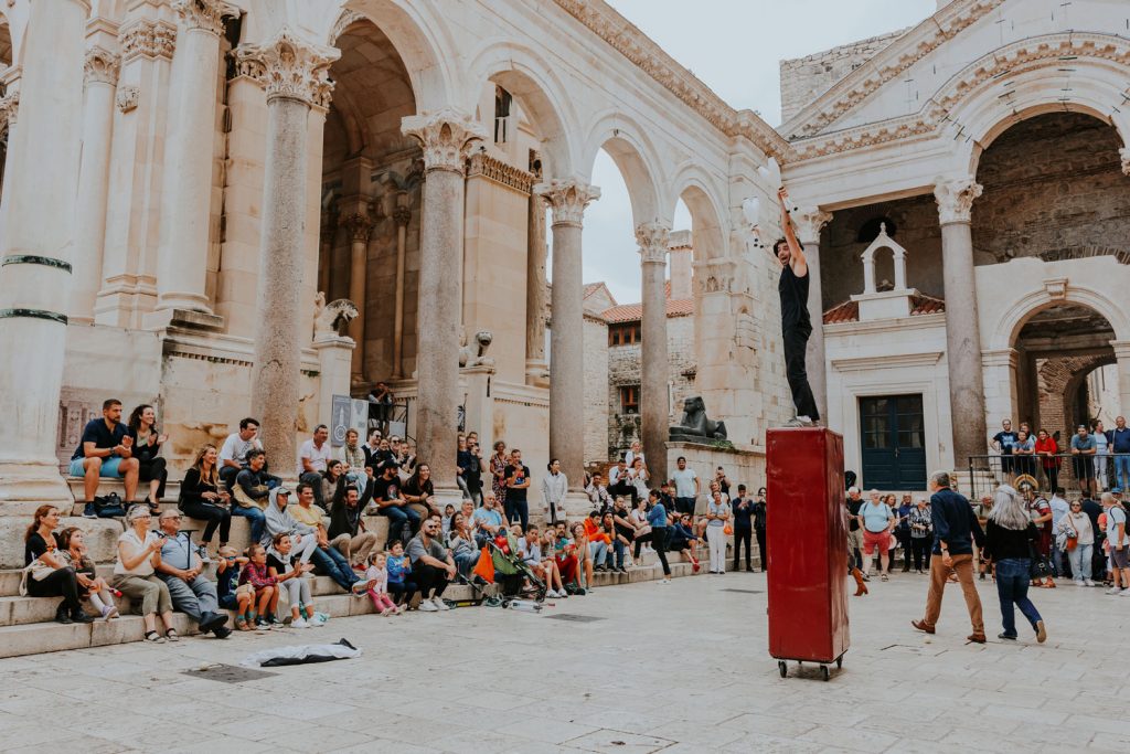 Affiche du spectacle Niente Panico ! de la cie Garuskji. Un homme debout sur un casier en métal sur une place de style italien.