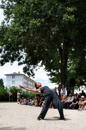 Une danseuse debout, tenue noire, cambrée et bassin en avant est entourée du public.