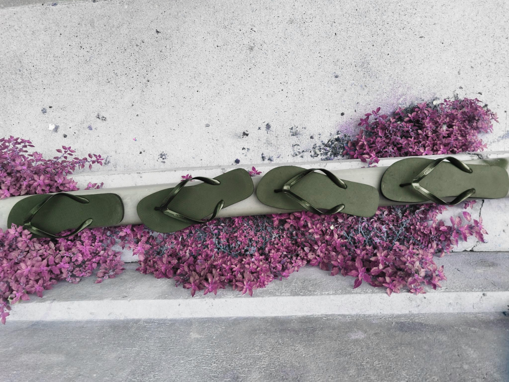 Arête de trottoir sur lequel sur disposé des fleurs roses et aligné des tongs noires.