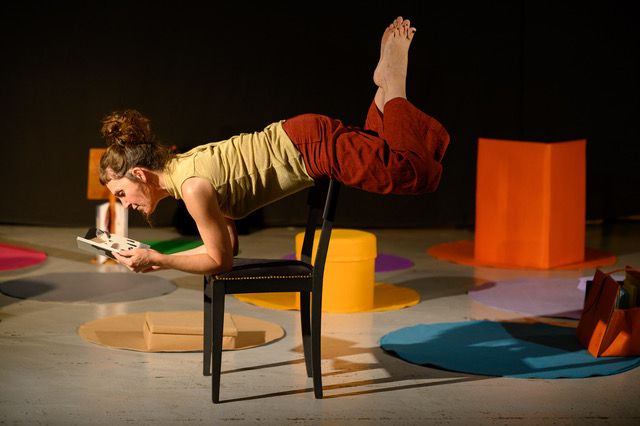 Une femme sur le ventre en équilibre sur une chaise lit un livre. Des objets de couleurs sont présents autour d'elle.