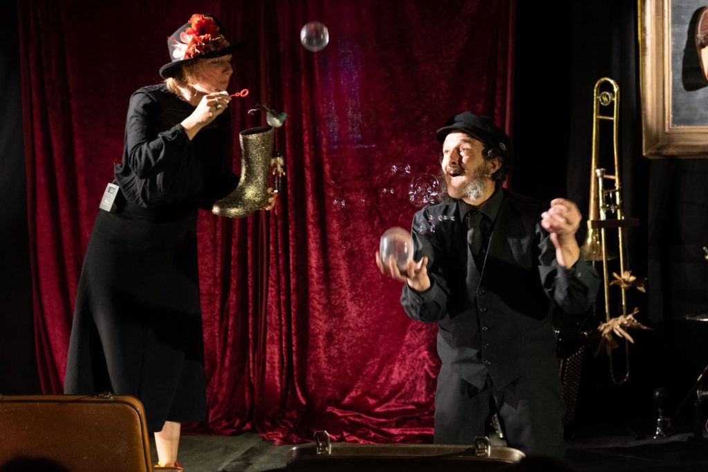 Sur scène, un homme et une femme en costumes noirs jonglent et font des bulles.