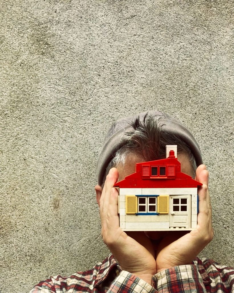 Un homme en bonnet se cache la tête avec une maison miniature qu'il tient dans ses mains.