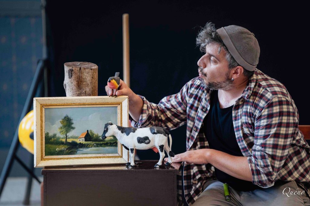 Un homme en bonnet et chemine à carreaux entasse des objets à côté de lui. Peintures, animaux en plastiques...
