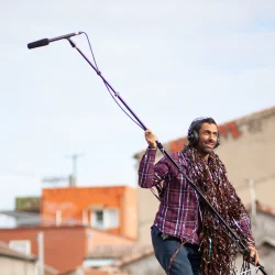 Un homme avance, casque sur la tête et micro perche dans les bras en extérieur pour écouter les sons qui l'entourent.
